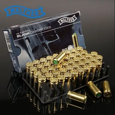 A Malther brand box of 9 mm PAK blank cartridges displayed next to a black tray holding multiple brass-cased cartridges with green tips, on a dark reflective surface.